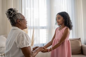 An older woman smiles as she holds her granddaughter’s hands, appreciating grandkids helping older adults.