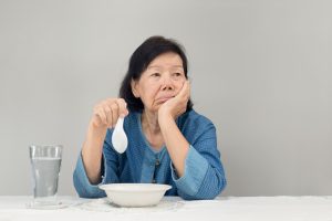 An older woman looking lonely and sad while trying to eat a bowl of food.