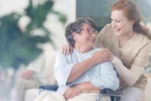 smiling senior woman in wheelchair looking over her should at smiling caregiver leaning in