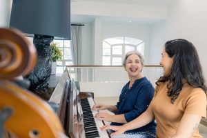 Empowering You to Live Well A woman receiving dementia care in Burlington smiles brightly at her caregiver as they play the piano together.
