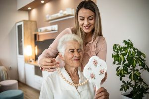 An older woman receiving personal care services smiles in the mirror as her caregiver places a strand of pearls around her neck.