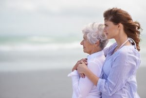 A caregiver handling the responsibilities of caregiving hugs the senior she cares for as they look out on the beach.