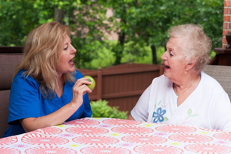 An older woman practices speech therapy exercises with her therapist at home.