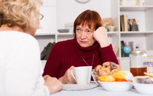 A woman needing support for family caregivers looks sternly at her sister over a cup of coffee.